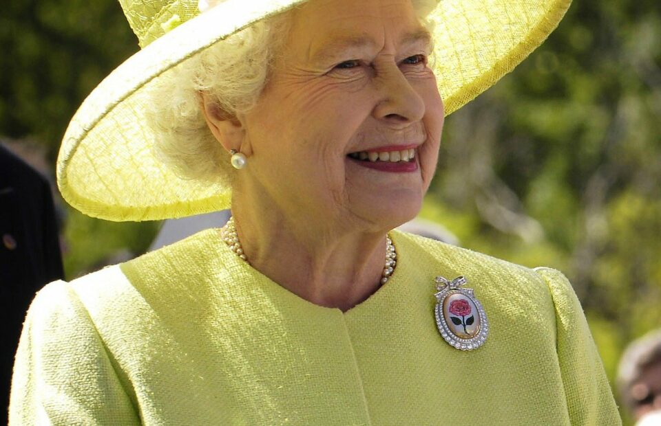 Queen Elizabeth II of England is wearing a yellow hat with flowers and a yellow jacket with a round brooch. She is smiling.
