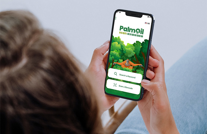 Woman holding iphone in hand showing home screen of PalmOil Scan App showing a cartoon orangutan amongst trees. Text reads "PalmOil scan"
