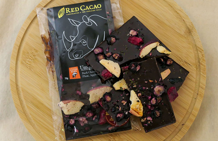 Dark chocolate block with freeze dried raspberries, apple and mint broken into pieces lying on top of dark chocolate block wrapped in clear packaging, resting on round wooden chopping board. Text on packaging reads 'Red Cacao Umqali' with Monarto Safari Park logo.