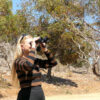 Woman with long blonde hair wearing black and brown striped top looking through black binoculars in bushland
