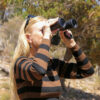 Woman with long blonde hair wearing black and brown striped top looking through black binoculars in bushland